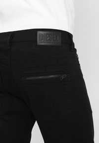 Pantalones de mezclilla negros con una etiqueta de cuero que dice "DIESEL", con un bolsillo con cremallera en la parte trasera y tejido texturizado.