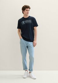 Tamsiai mėlynas medvilninis marškinėlis trumpomis rankovėmis su baltu grafiniu tekstu. Sukomplektuota su šviesiai mėlynomis džinsais ir baltais sportbačiais.