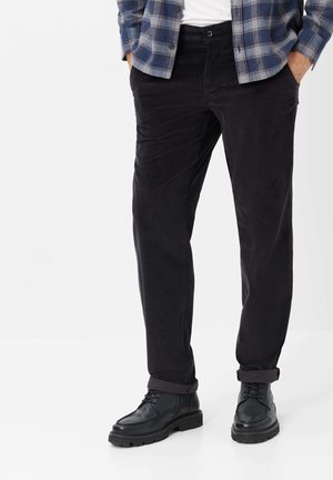 Uomo che indossa pantaloni in velluto a coste neri con risvolti, stivali neri con lacci, una camicia bianca e una camicia a quadri blu con bottoni. Mani nelle tasche.