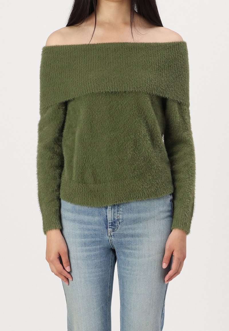 Femme portant un pull vert duveteux à épaules dénudées et un jean bleu clair, debout devant un fond clair uni.