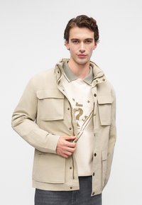 Šviesiai ruda striukė su užtrauktuku priekyje, snap užtrauktukais ir keliais kišenėmis. Dėvima ant šviesios spalvos marškinėlių su žaliu apykakle. Lygus paviršius.