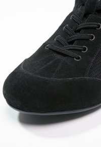 Närbild av en svart mockasneaker med svart snörning och meshsidopanel mot en vit bakgrund.