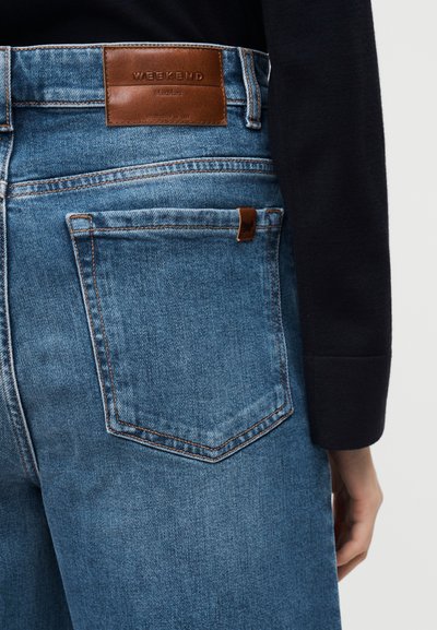 Μπλε τζιν παντελόνι με μεγάλο πίσω τσέπη και καφέ δερμάτινο σημάδι με την ένδειξη "WEEKEND", φορεμένο με μαύρο μακρυμάνικο πάνω μέρος που καλύπτει τον βραχίονα.