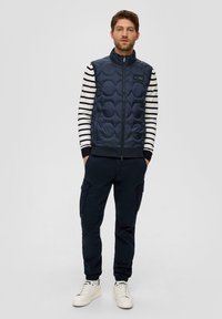 Marineblau gestepptes Westchen über einem weiß-schwarz gestreiften Langarmshirt, kombiniert mit dunklen Cargo-Hosen und weißen Sneakers.