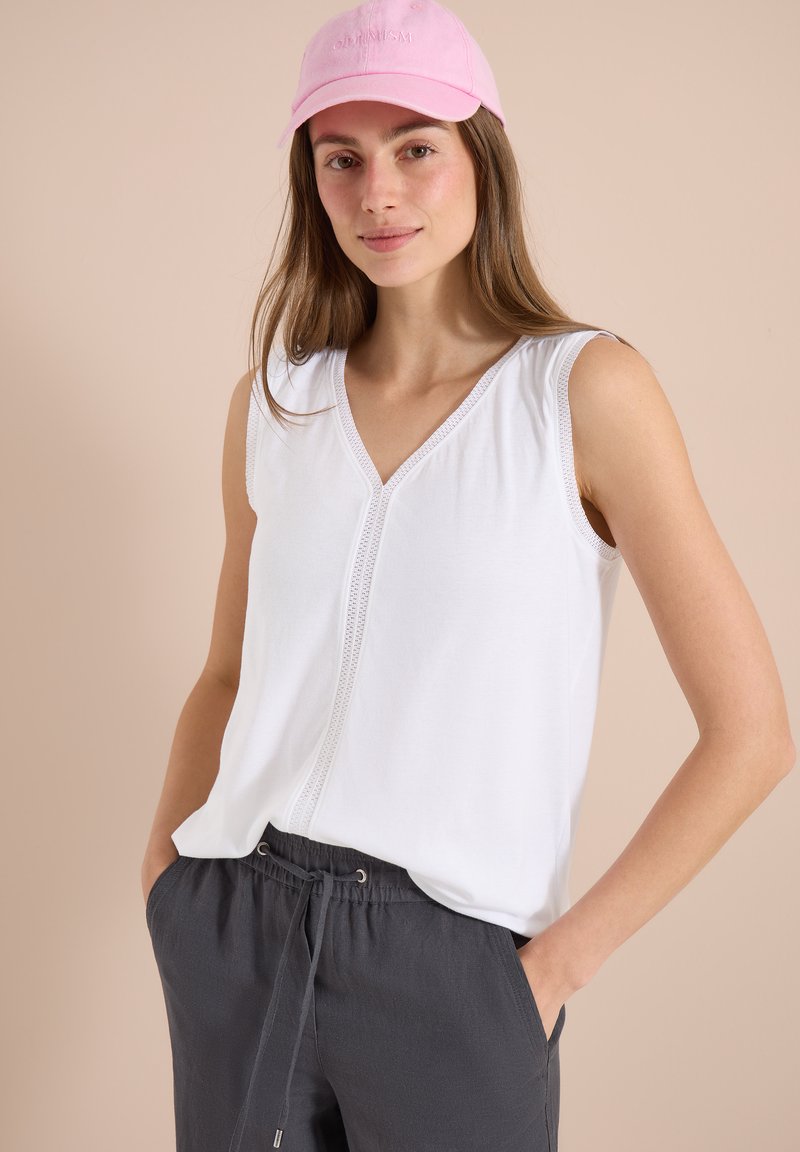 Femme portant une casquette rose, un haut blanc sans manches en V avec bordure en dentelle, et un pantalon ample gris foncé à cordon, debout, les mains dans les poches.