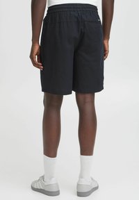 Shorts de sport noirs avec une taille élastique, coutures visibles et une poche latérale. Portés avec des baskets grises et des chaussettes blanches.