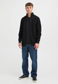 Schwarze Reißverschluss-Strickjacke aus strukturiertem Stoff, getragen über einem schwarzen Shirt. Kombiniert mit blauen Jeans und schwarzen Sneakers. Stehende Haltung, neutraler Hintergrund.