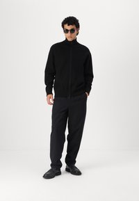 Maglione nero con zip realizzato in materiale a maglia, abbinato a pantaloni scuri e scarpe nere. Presenta un colletto alto e polsini a coste.