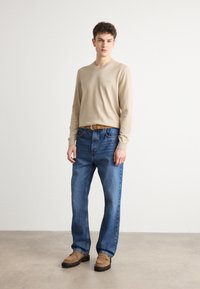 Beige, hosszú ujjú pulóver v-kivágással, kék egyenes szabású farmernadrággal párosítva. A barna öv és a laza barna cipők teszik teljessé az öltözetet.