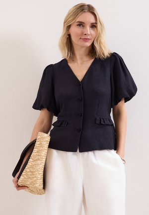 Vrouw met schouderlang blond haar, draagt een zwart blousje met knopen en pofmouwen en een witte broek, houdt een gevlochten strohandtas vast.