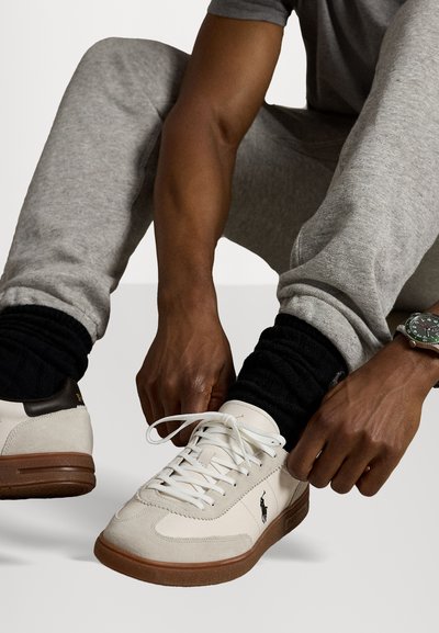 Bijele i bež suede tenisice s smešnim gumama. Značajke uključuju bijele vezice i logo sa strane. Model vezuje cipele.