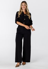 Vrouw met lang blond haar, gekleed in een zwarte overhemd met knopen, zwarte wijde pantalons, gouden armbanden en leopardprint hakken, staand op een witte achtergrond.