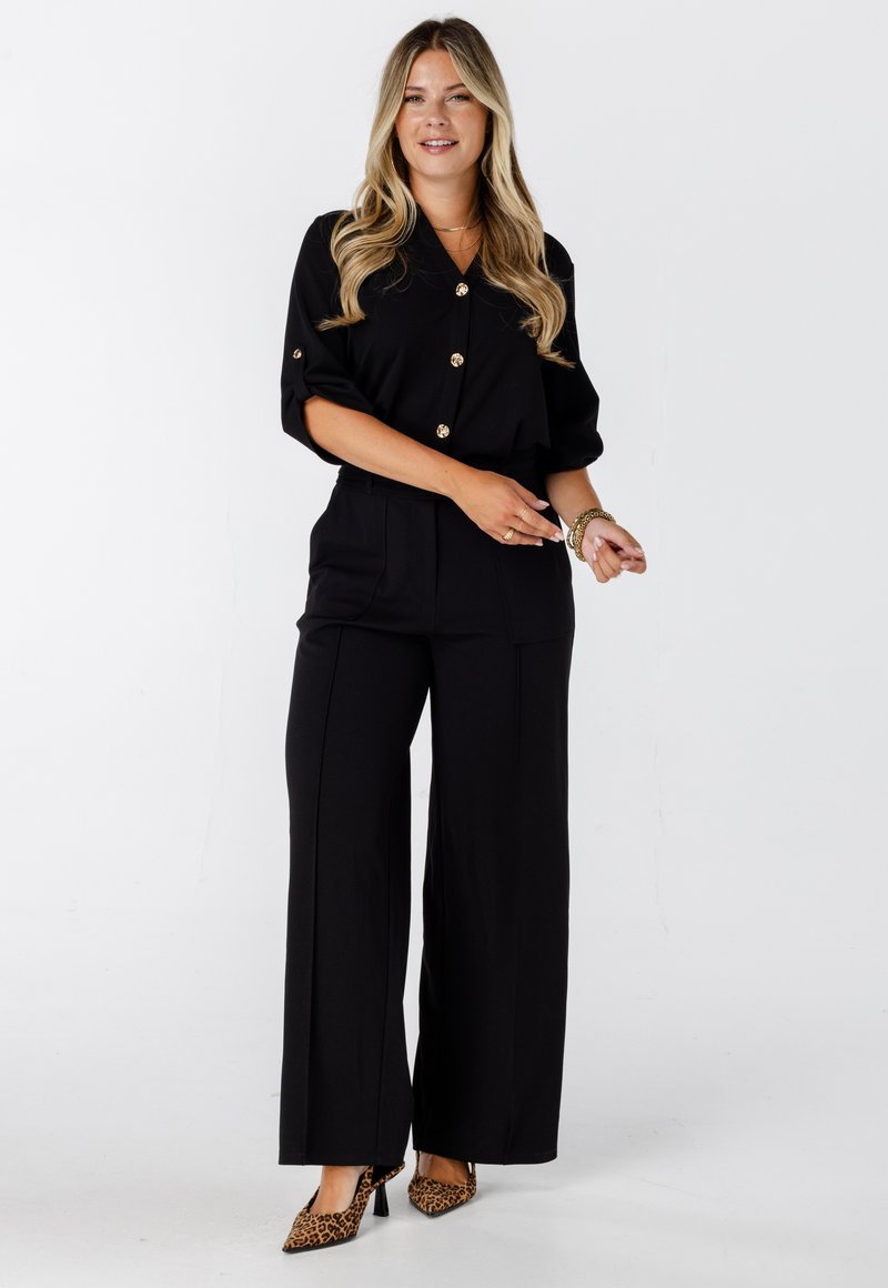 Vrouw met lang blond haar, gekleed in een zwarte overhemd met knopen, zwarte wijde pantalons, gouden armbanden en leopardprint hakken, staand op een witte achtergrond.