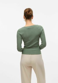 Grön ribbad långärmad topp med en kroppsnära silhuett, ihop med beiga vida byxor. Bakgrunden är helt vit.