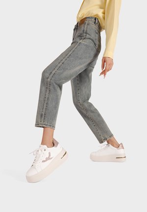 Persona con jeans de lavado claro y zapatillas de plataforma blancas con detalles de purpurina, levantando una pierna mientras está de pie contra un fondo liso.