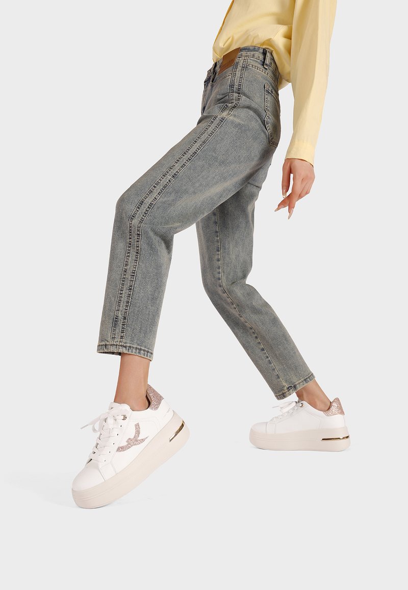 Persona che indossa jeans chiari e sneakers bianche con dettagli glitterati, sollevando una gamba mentre si appoggia a uno sfondo semplice.