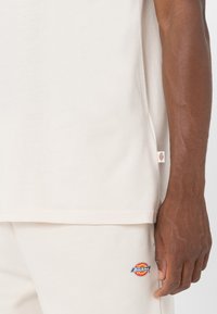 Camiseta de algodón beige con un corte relajado, que presenta un pequeño parche con el logo de Dickies en el lado y manga corta.