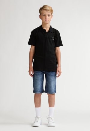 Zwart poloshirt met korte mouwen, gecombineerd met blauwe spijkershorts. Witte sneakers en witte enkelsokken maken de outfit compleet.