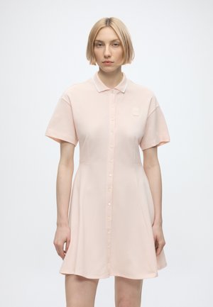 Femme aux cheveux blonds courts portant une robe rose clair à manches courtes boutonnée avec col, debout devant un fond blanc uni.