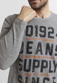 Grå langærmet t-shirt med store sorte og orange bogstaver, med rund hals og blødt, let struktureret stof.