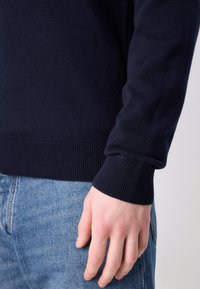 Pull bleu marine foncé avec des poignets côtelés, texture douce et design ajusté, associé à un jean en denim bleu clair. Main visible reposant sur le denim.