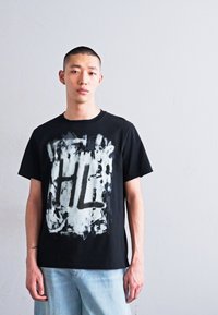 Fekete pamut póló fehér és szürke absztrakt grafikai mintával, amelyen az "HL" betűk láthatók. A modellen világoskék farmernadrág van.