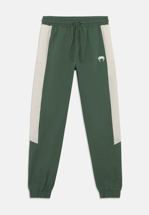Green jogger pants with cream side panels, elastic waistband, drawstring, and ribbed cuffs; features a small white logo on the front.