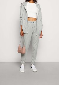 Woman wearing light gray zip-up hoodie and sweatpants with white crop top, white Nike sneakers, holding small pink handbag with chain strap.