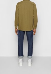 Oliwkowa koszula z długim rękawem z zaokrąglonym dołem i zapinanymi rękawami, w połączeniu z ciemnoniebieskimi dżinsami z mankietem i białymi butami.