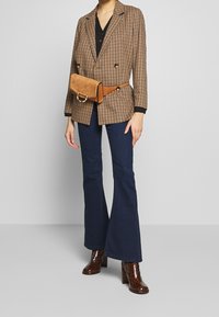 Blazer marron à carreaux avec des bordures noires, associé à un jean foncé évasé et des bottes à talons brillantes marron. Un sac banane en suède beige est porté à la taille.