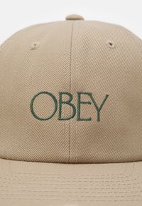 Boné de beisebol caqui com bordado verde "OBEY", tecido texturizado, construção de seis painéis e coroa arredondada.
