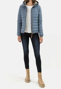 Lysblå pufferjakke med hætte, lynlås foran og tekstureret stof. Båret med en hvid top og mørke jeans, suppleret med lysebrune ankelstøvler.