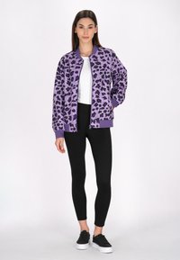 Giacca bomber viola con stampa animale nera, con polsini e orlo a coste, indossata sopra una camicia bianca e leggings neri con sneakers nere.