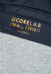 Metka z bawełnianej tkaniny na ubraniu, z granatowymi i szarymi sekcjami. Żółty tekst przedstawia "JJ CORE L.A.B." oraz "CTM No. - 72030111 < 1975 >."