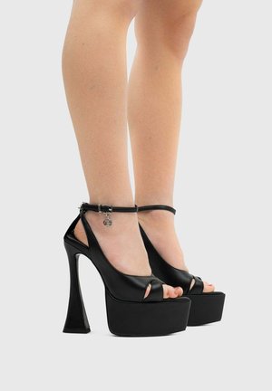 Gambe che indossano décolleté nere con plateau, cinturini alla caviglia e punta aperta, mostrate su uno sfondo chiaro uniforme.