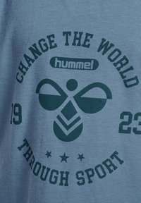 Ljusblå t-shirt med mörkgrön tryckt text och logotyp. Innehåller "ÄNDRAR VÄRLDEN GENOM SPORT" och "hummel", med grafiska element.