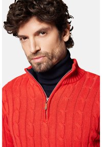 Czerwony sweter z dzianiny w warkocze z pół zamkiem i wysokim kołnierzem, noszony na granatowym golfie. Miękka faktura z prążkowanym wzorem.