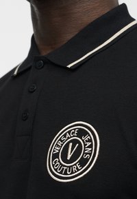 Μαύρο πόλο μπλουζάκι με λευκή διπλή ρίγα στον γιακά και κυκλικό κεντημένο λογότυπο Versace Jeans Couture στο στήθος.