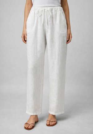 Pantaloni larghi in lino bianco con vita elastica e coulisse indossati con sandali piatti color cuoio su una persona in piedi.