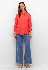 Donna che indossa una blouse a bottoni corallo brillante, jeans a gamba larga blu e stivaletti in suede beige, in piedi contro un muro bianco.