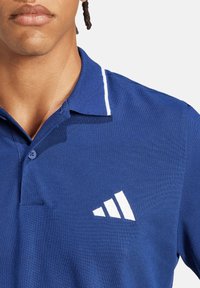Polo blu in tessuto texturizzato, con logo a tre strisce bianco sul petto sinistro e bordo bianco a contrasto sul colletto.
