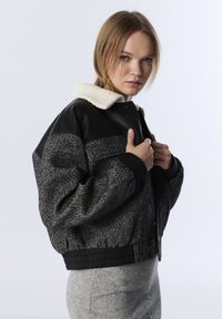 Jonge vrouw met licht haar, gekleed in een zwart en grijs wollen jasje met leren details en een witte kraag, die de voorkant van het jasje aanpast.