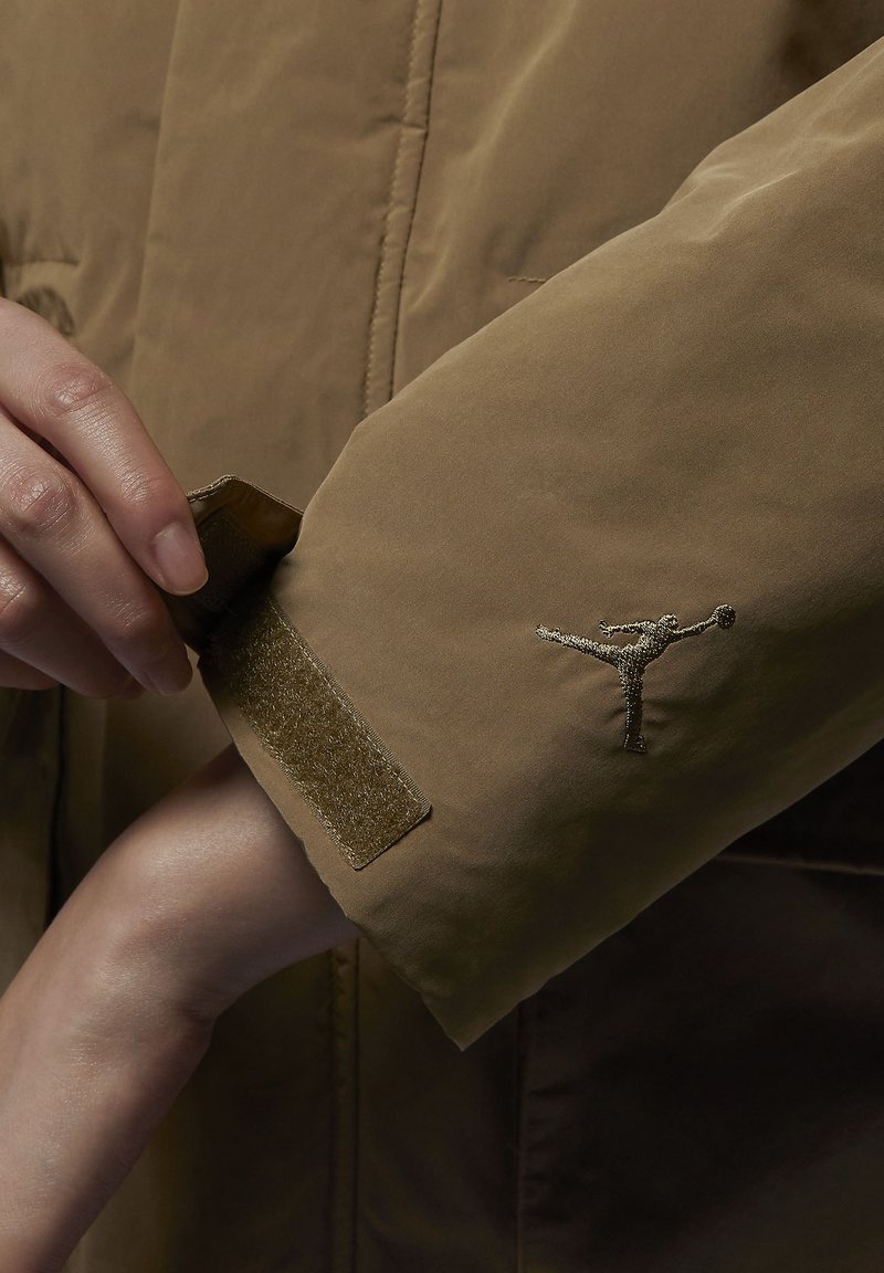 Chaqueta impermeable marrón con cierre de muñeca de velcro y un logo bordado en plata en la manga. Textura suave con un diseño elegante.