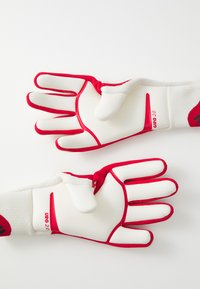 Gants de gardien de but blancs avec des accents rouges. Les caractéristiques incluent des épines au niveau des doigts, une surface de prise texturée et des sangles élastiques au poignet. Modèle étiqueté URG 2.0.