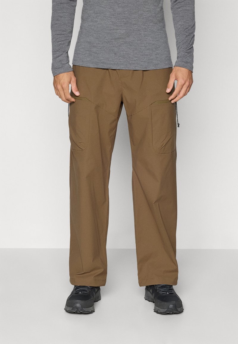 Pantalons de plein air marron avec une coupe décontractée, poches cargo et zippers. Le matériau est léger et texturé, associé à des chaussures noires.