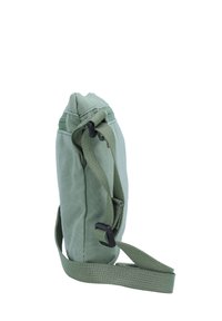 Saco de lona verde com uma textura suave, design slim, alça ajustável e detalhes em metal preto. A vista lateral mostra a forma compacta e as costuras.
