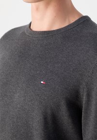 Gros plan d'une personne portant un pull en maille gris foncé avec un petit logo rectangulaire rouge, blanc et bleu sur la poitrine.