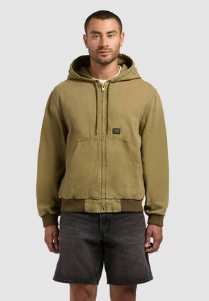 Man wearing a tan zip-up hooded jacket with front pockets and dark gray knee-length shorts against a plain background.