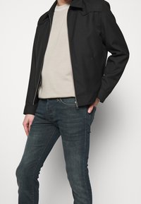 Svart zipjacka med hög krage, bärs över en ljusbeige skjorta, tillsammans med mörkblå jeans. Jackan har en strukturerad design.