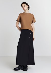 Top corto marrone a maniche corte abbinato a una gonna avvolgente nera con accenti circolari metallici. L'outfit è completato da stivaletti neri alla caviglia.